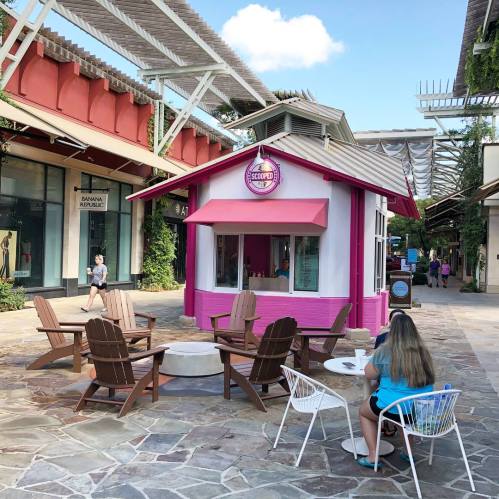 A colorful ice cream stand with outdoor seating in a shopping area, surrounded by trees and shops.