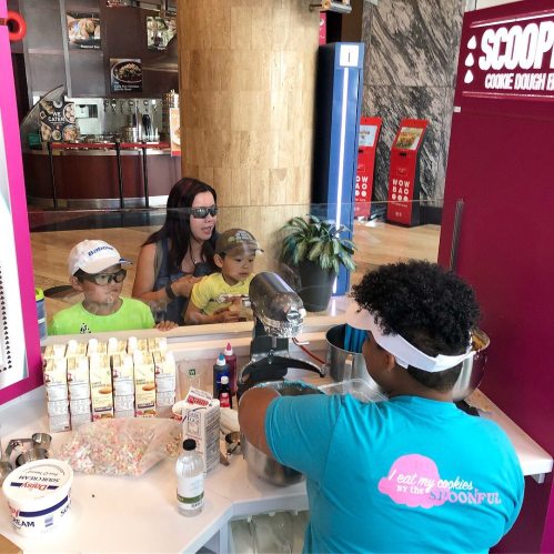 A woman and two children watch as a worker prepares cookie dough at a colorful dessert shop.