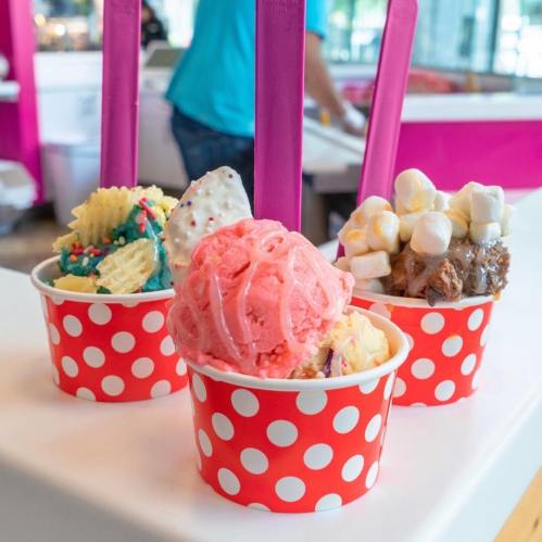 Three colorful ice cream cups with vibrant toppings, served in red polka dot containers, with purple spoons.