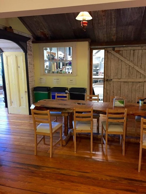 A rustic dining area with a wooden table, chairs, and a mirror, featuring a corrugated metal ceiling and wooden floors.