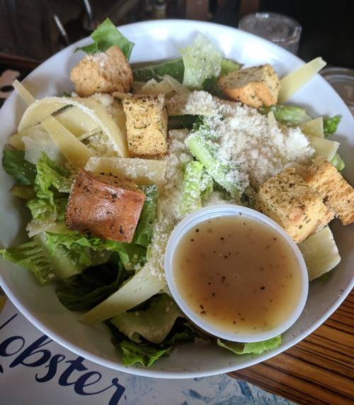 A fresh salad with romaine lettuce, croutons, Parmesan cheese, and dressing in a cup, served in a white bowl.