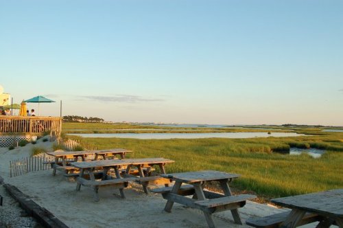 A serene beach scene with picnic tables, marshland, and a calm waterway under a clear sky.