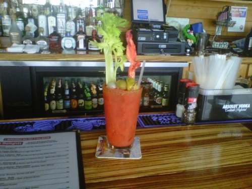 A vibrant Bloody Mary cocktail garnished with celery, olives, and shrimp, sitting on a bar counter.