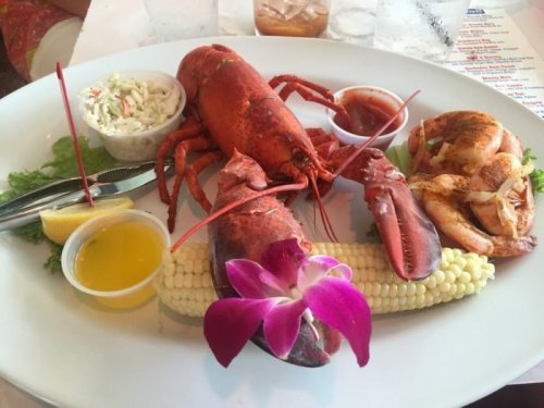 A plate featuring a whole lobster, shrimp, corn, coleslaw, and garnished with a flower and lemon.