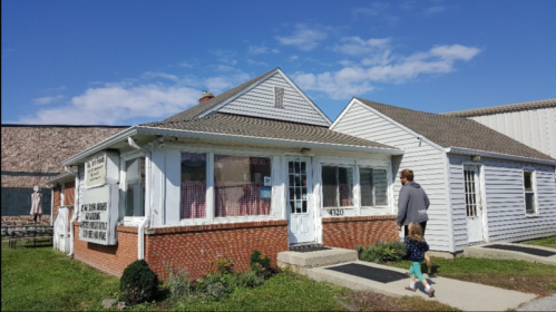 A person walks past a small, brick-front building with a sign, under a clear blue sky.