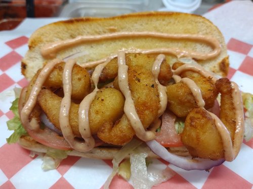 A sandwich with fried shrimp, lettuce, tomato, and drizzled sauce on a toasted bun, served on a checkered paper.