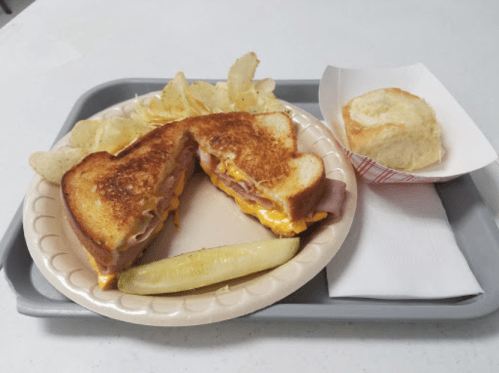 A plate with a grilled cheese sandwich, chips, a pickle, and a biscuit on a tray.