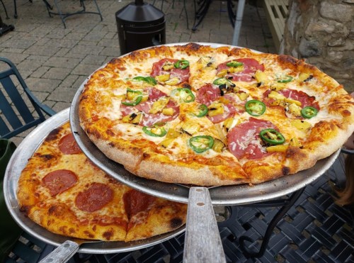 Two pizzas on metal trays: one topped with pepperoni, the other with various toppings including jalapeños and pineapple.