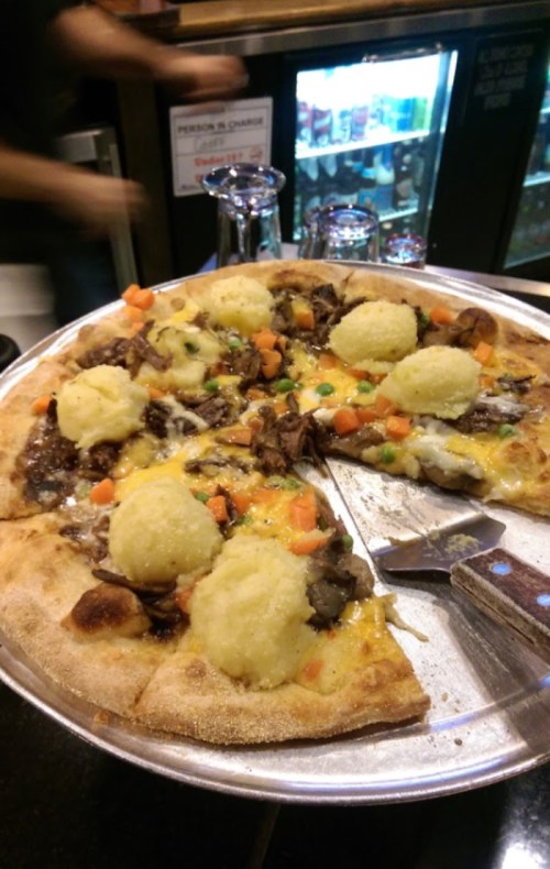 A pizza topped with shredded beef, cheese, and dollops of mashed potatoes, served on a metal tray.
