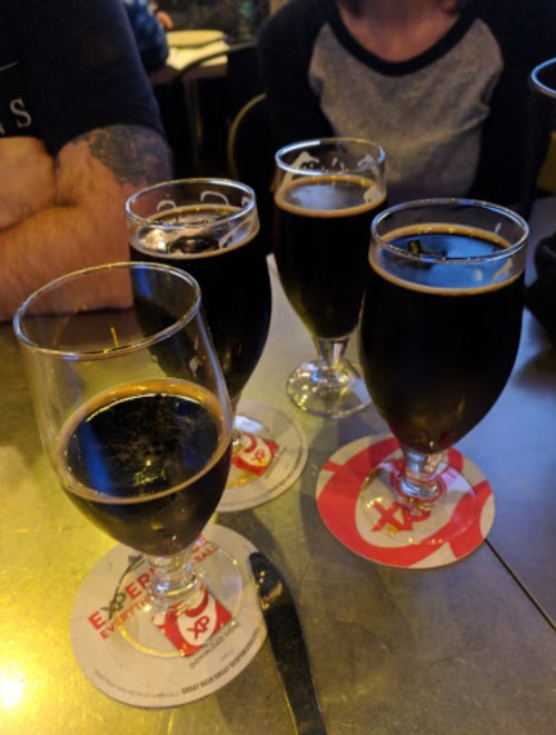 Four glasses of dark beer on a table with coasters, with a blurred background of people and a restaurant setting.