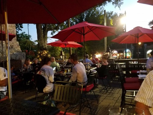 Outdoor dining scene with red umbrellas, tables filled with people enjoying food and drinks in a lively atmosphere.