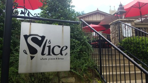 Sign for "Slice" restaurant with outdoor seating and red umbrellas, surrounded by greenery and stairs leading up.