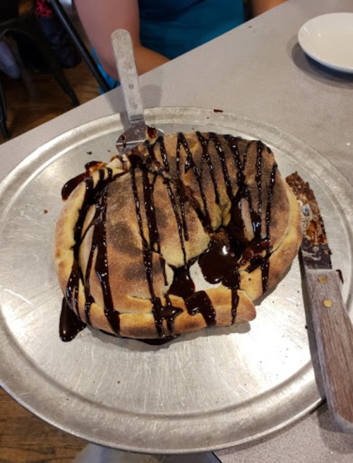 A large, warm dessert with a chocolate drizzle on a silver platter, partially cut with a knife beside it.