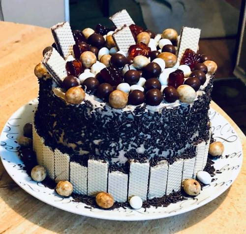 A round chocolate cake topped with wafers, chocolate balls, and colorful candies, displayed on a decorative plate.