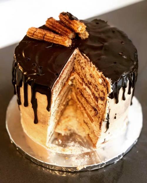 A layered cake with chocolate ganache dripping down, topped with churros, and partially sliced to reveal its layers.