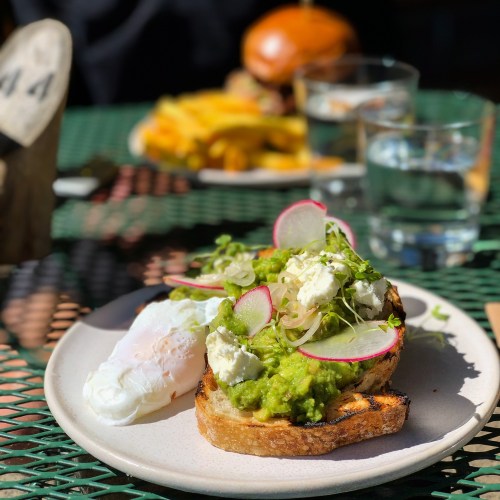 A plate with avocado toast topped with radishes and feta, alongside a poached egg, with a burger and fries in the background.