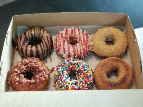 A box of six assorted donuts: chocolate glazed, strawberry drizzled, coconut, sprinkles, and two others with unique toppings.