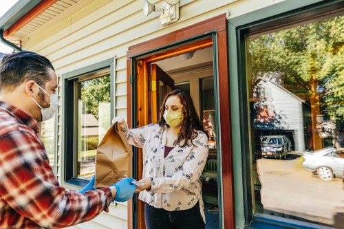 A person in a mask hands a brown paper bag to another person at a doorway, both wearing gloves.