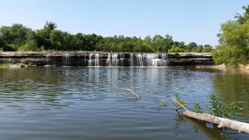 A serene waterfall cascading over rocky ledges into a calm lake, surrounded by lush greenery under a clear blue sky.
