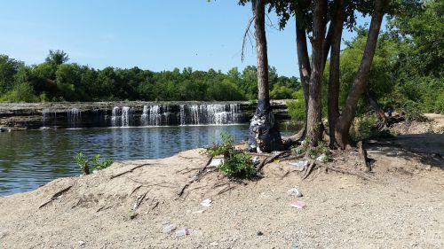 A serene waterfall cascades into a calm river, surrounded by trees and litter on the sandy shore.