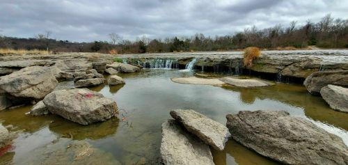 A serene landscape featuring a small waterfall over rocky terrain, surrounded by calm water and overcast skies.