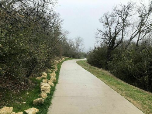 A winding concrete path surrounded by trees and shrubs on a cloudy day.