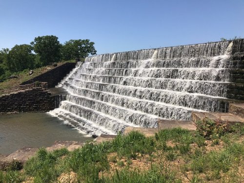 A cascading waterfall flows over a stepped stone structure, surrounded by greenery and a clear blue sky.
