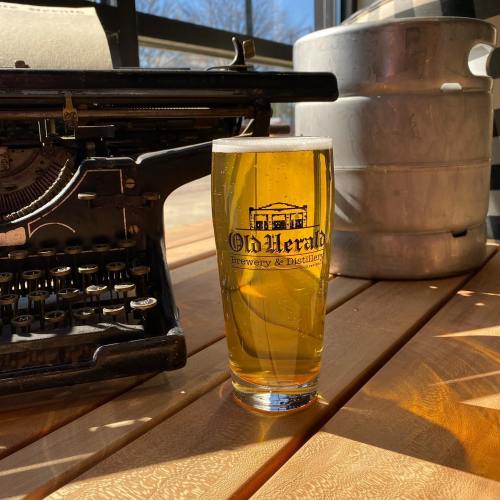 A glass of golden beer sits on a wooden table next to a vintage typewriter and a keg in a bright setting.