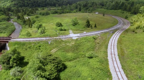 Aerial view of a winding railway track through lush green fields and trees, with a river nearby.