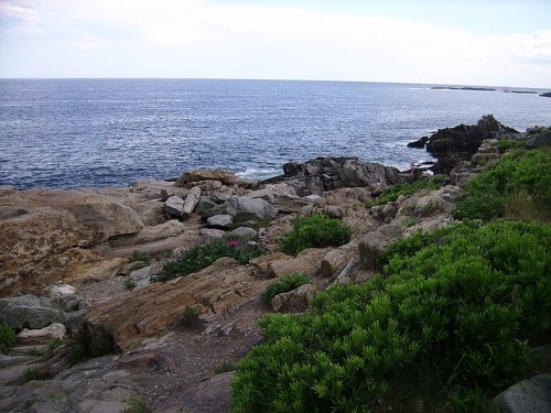 Rocky shoreline with green vegetation and calm blue ocean under a partly cloudy sky.