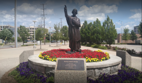 Statue of Bishop Francis Asbury surrounded by colorful flowers and greenery, with a blue sky in the background.