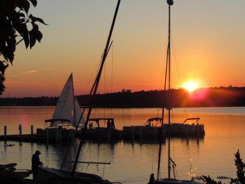 A serene sunset over a calm lake, with sailboats docked and silhouettes of masts against the colorful sky.