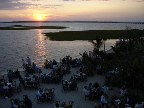 A sunset over a calm waterway, with diners seated at tables on the beach, surrounded by palm trees.