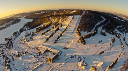 Aerial view of a snowy landscape with ski slopes, trees, and buildings under a clear sky during sunset.