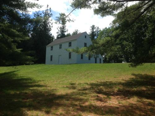 A green, two-story house sits on a grassy hill, surrounded by trees under a partly cloudy sky.