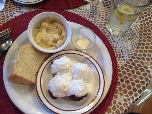 A plate with biscuits and gravy, mashed potatoes, a small cup of cream, a slice of bread, and a glass of water with lemon.