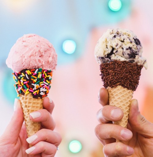 Two hands hold ice cream cones: one with pink ice cream and colorful sprinkles, the other with cookies and cream topped with chocolate sprinkles.