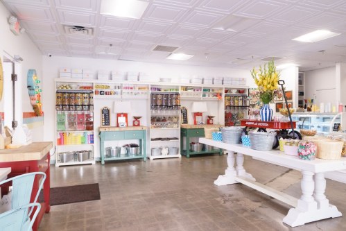 Brightly lit shop interior with colorful shelves, jars, and baskets, featuring a large table and decorative flowers.