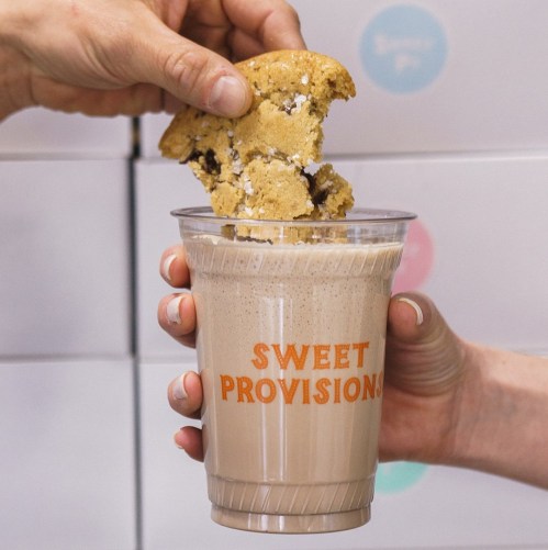 A hand dips a cookie into a milkshake in a cup labeled "SWEET PROVISIONS."