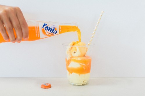 A hand pours orange Fanta over a scoop of ice cream in a clear cup, with a straw and bottle cap nearby.