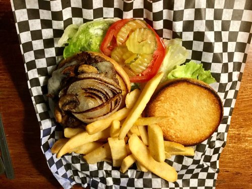 A burger with melted cheese, grilled onions, lettuce, tomato, pickles, and fries on a checkered paper.
