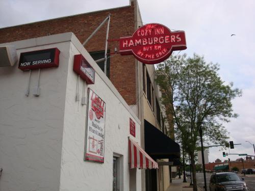 A corner view of the Cozy Inn Hamburgers restaurant, featuring a red sign and outdoor seating area.