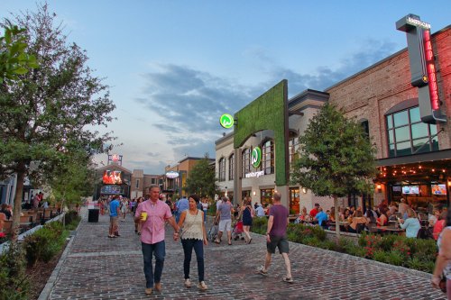 A lively outdoor scene with people walking, dining, and enjoying a vibrant atmosphere at a restaurant area.