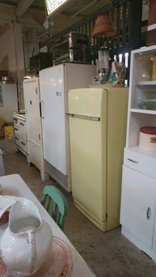A row of vintage refrigerators and appliances in a retro-style setting, featuring a yellow fridge and a white cabinet.