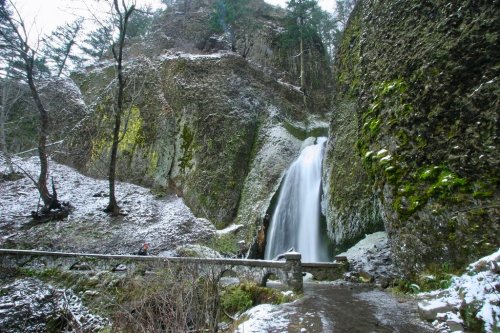 A serene waterfall cascades down rocky cliffs, surrounded by snow-dusted trees and mossy stones in a tranquil forest setting.