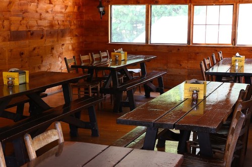 Interior of a rustic wooden dining area with long tables, chairs, and large windows letting in natural light.