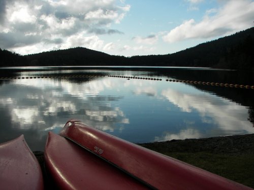 Two red kayaks rest on the shore, reflecting clouds and mountains on a calm lake under a partly cloudy sky.