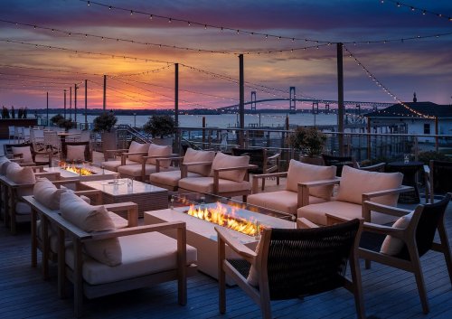 A cozy outdoor seating area with a fire pit, overlooking a sunset and a bridge, adorned with string lights.