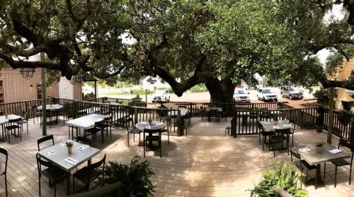 Outdoor restaurant patio with wooden tables and chairs, surrounded by greenery and large trees. Cars parked nearby.