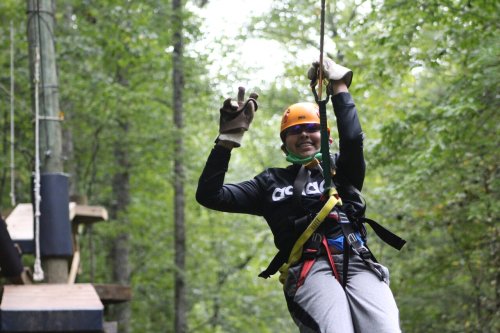 A person in a helmet and gloves smiles while zip-lining through a forested area.